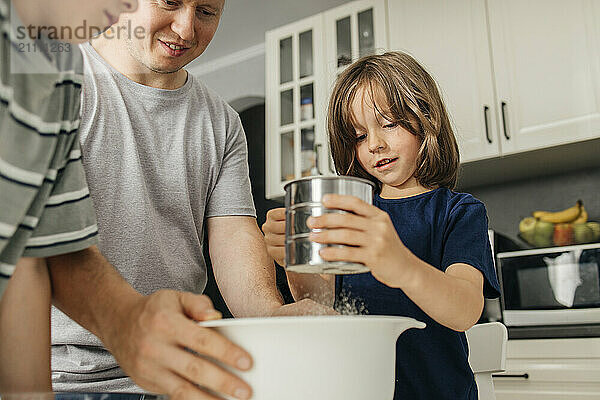 Cute boy sifting flour with father and brother in kitchen