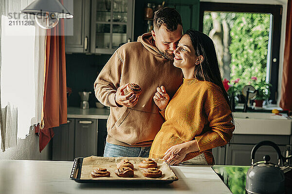 Affectionate man holding cinnamon bun with pregnant wife in kitchen