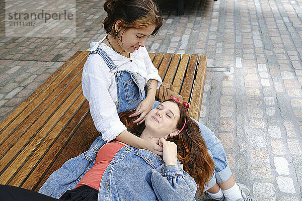 Young woman lying on lap of girlfriend sitting on bench at sidewalk