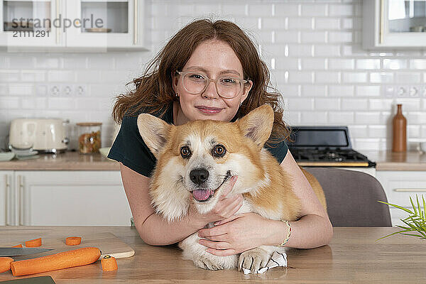 Smiling woman embracing Welsh Corgi dog in kitchen at home