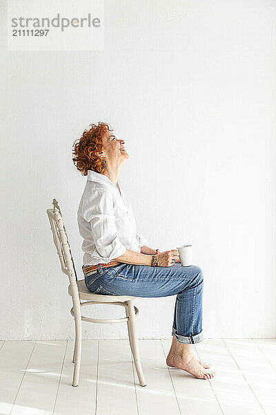 Smiling woman holding coffee mug and sitting on chair in front of white wall