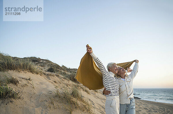 Happy mother and daughter holding shawl and enjoying leisure time at beach