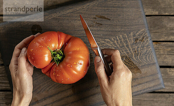 The hands of a woman with a kitchen knife on a wooden cutting board with a red tomato.