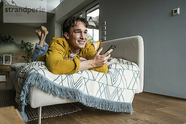 Thoughtful young man lying on sofa and holding smart phone at home