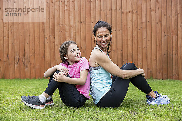 Mother and daughter sitting back to back on grass in back yard