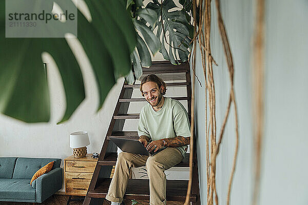 Smiling young freelancer sitting with laptop on staircase at home