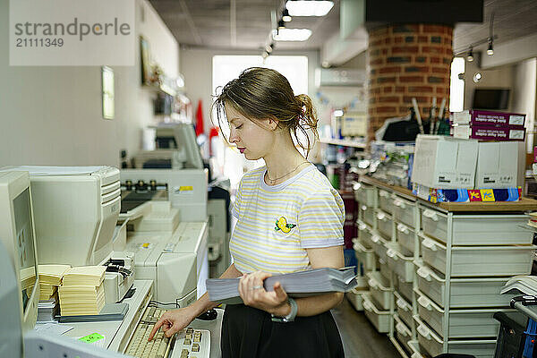 Woman holding stack of papers and using computer in a printing shop