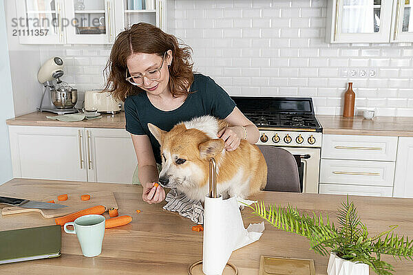 Young woman feeding carrot to dog in kitchen at home