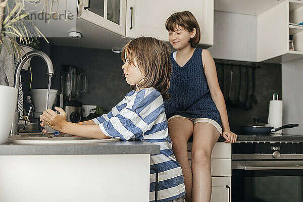 Smiling girl sitting and looking at brother washing dishes in kitchen