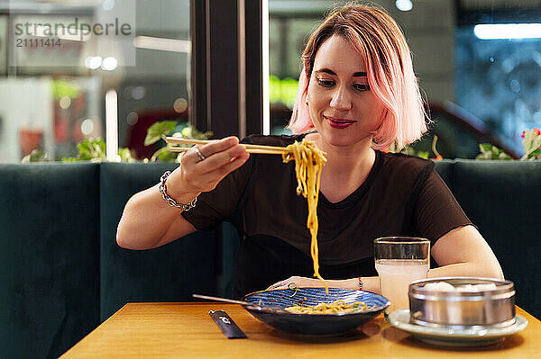 Smiling woman with pink hair eating noodles with chopsticks at restaurant