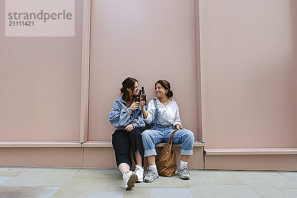 Happy gay couple toasting with beer bottles and sitting in front of pink wall