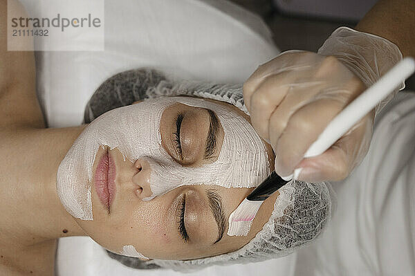 Hand of beautician applying face mask with brush on woman