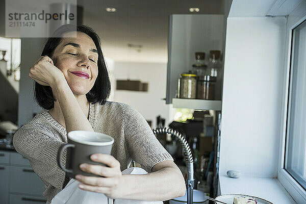 Smiling woman with hand on chin holding coffee cup at home