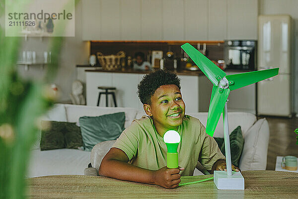 Happy boy powering light bulb through wind turbine model in living room at home