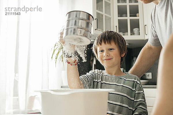 Smiling boy sifting flour with father in kitchen