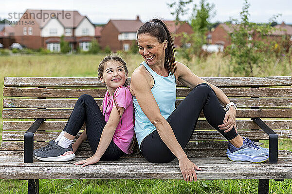 Happy woman sitting back to back with daughter on bench