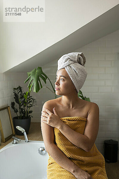 Thoughtful woman with towel wrapped around head sitting on bathtub in bathroom