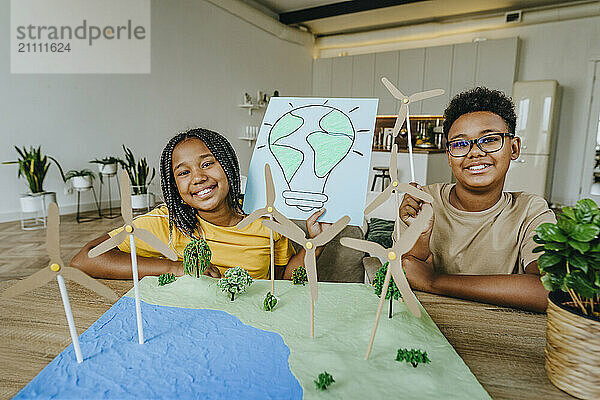 Happy siblings sitting with wind turbines project and light bulb drawing at table