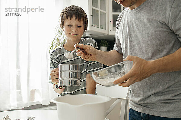 Smiling boy sifting flour and helping father in kitchen