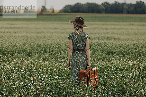 Woman wearing hat and holding suitcase standing in field