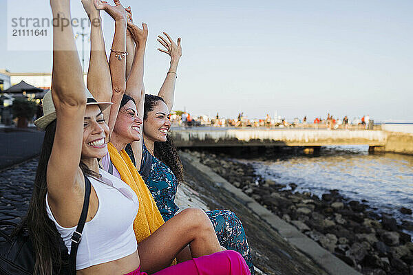 Cheerful friends with arms raised sitting on promenade