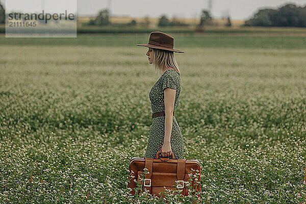 Blond woman holding suitcase and standing amidst field