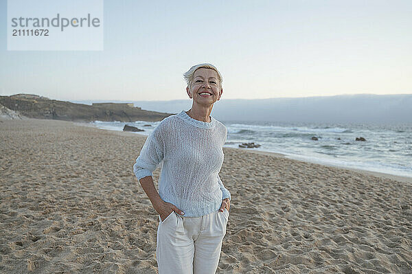 Smiling woman with hands in pockets standing at beach