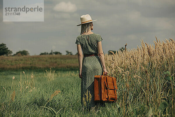 Blond woman holding suitcase and standing near reeds on sunny day