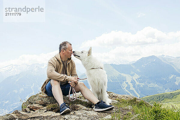 White Swiss Shepherd dog licking face of man sitting on rock by mountains