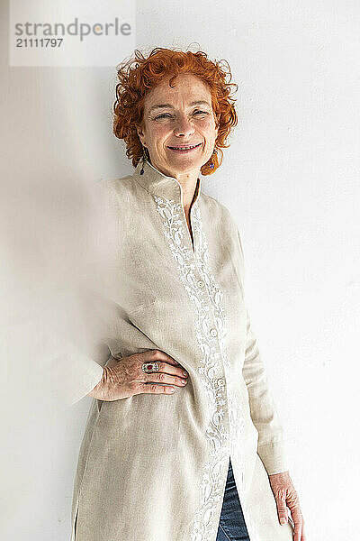 Smiling redhead woman with hand on hip and standing in front of white wall