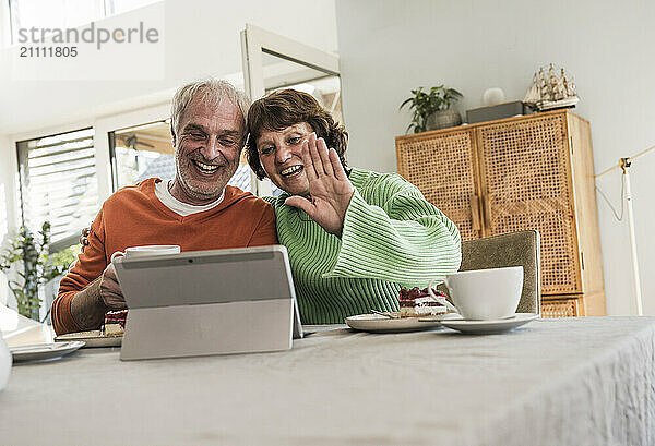 Cheerful couple talking on video call using tablet PC and sitting near table at home