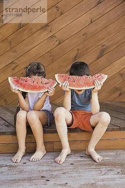 Boy and girl hiding faces with watermelon slices and sitting at porch