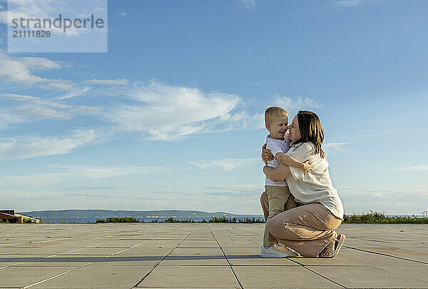 A dark-haired woman in a white shirt hugs her son against a blue sky.