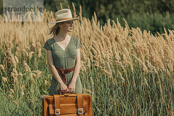 Beautiful woman holding suitcase and standing near reeds on sunny day