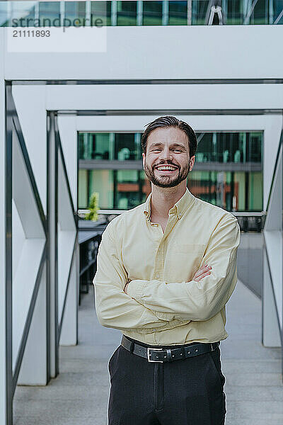 Smiling business person with arms crossed standing near office building entrance