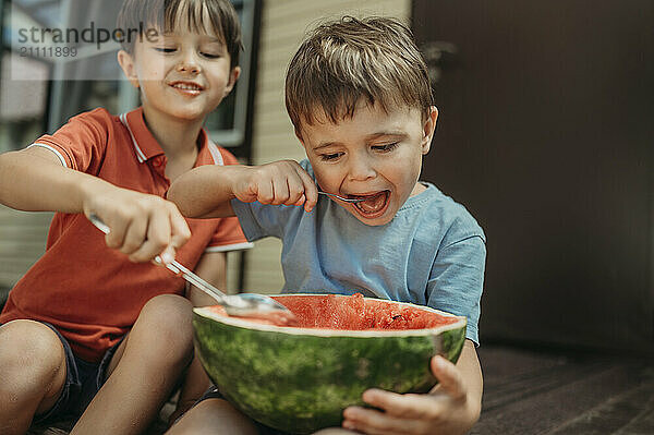 Brothers sitting and eating watermelon with spoon at backyard