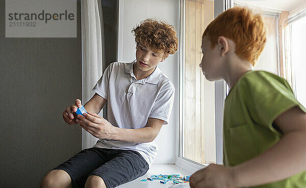 Boy holding toy block and sitting with brother on window sill at home