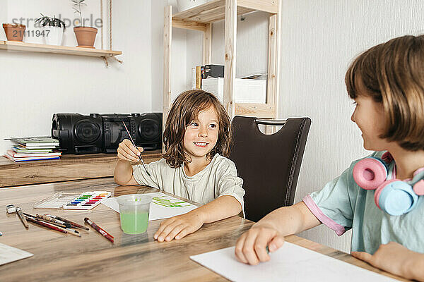 Cute boy holding paintbrush and sitting with sister near table at home