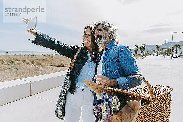 Smiling woman taking selfie with man carrying picnic basket