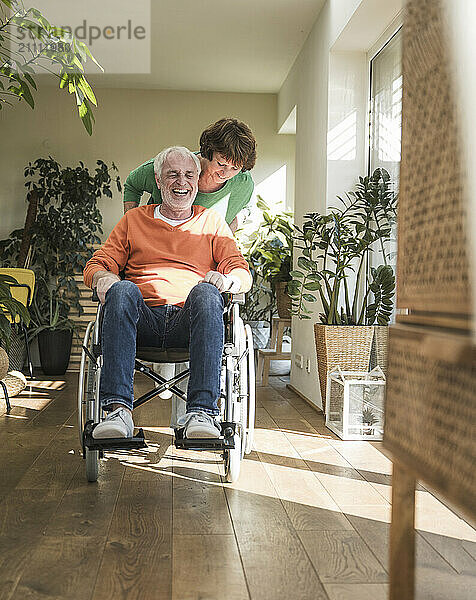 Happy senior man sitting on wheelchair pushed by wife at home