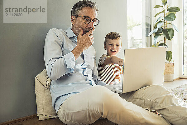 Man with hand on mouth using laptop by grandson at home