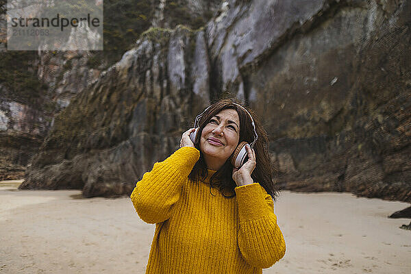 Smiling senior woman wearing wireless headphones and listening to music on beach