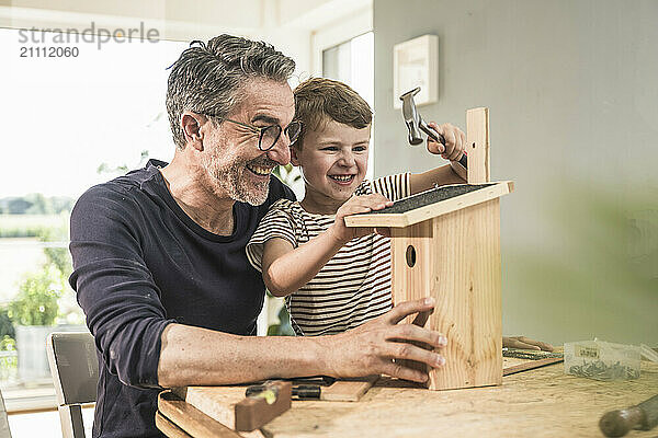 Cheerful boy timbering birdhouse by grandfather at home