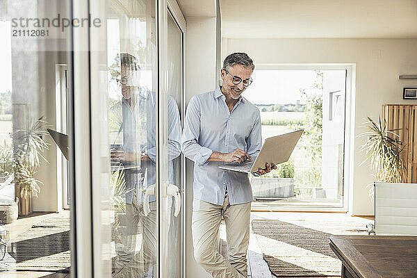 Smiling businessman using laptop leaning on window at home