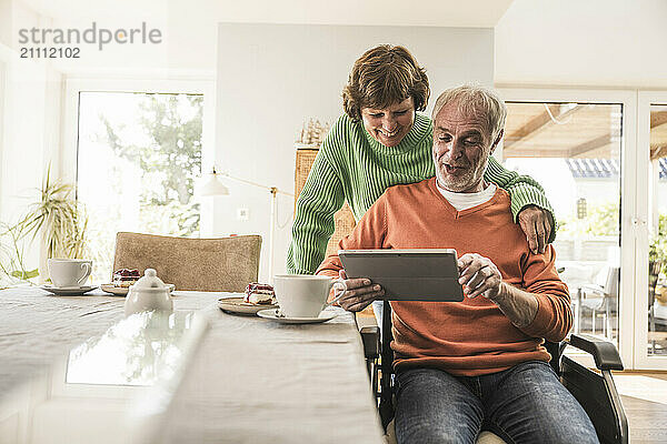 Happy senior woman with husband on wheelchair using tablet PC at home