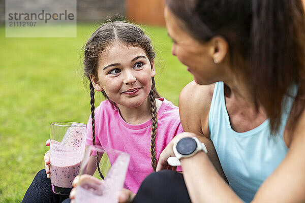 Girl with braided hair holding smoothie glass sitting next to mother in back yard