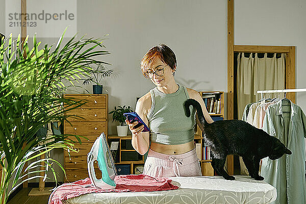 Woman using smart phone with black cat on ironing board at home