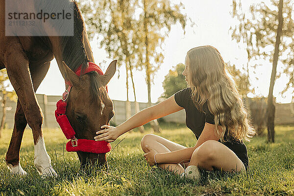 Girl sitting cross legged on grass with horse in farm