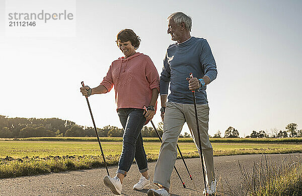 Senior man and woman walking with hiking poles on road near meadow