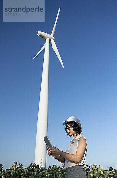 Young engineer using tablet PC standing near wind turbine at farm